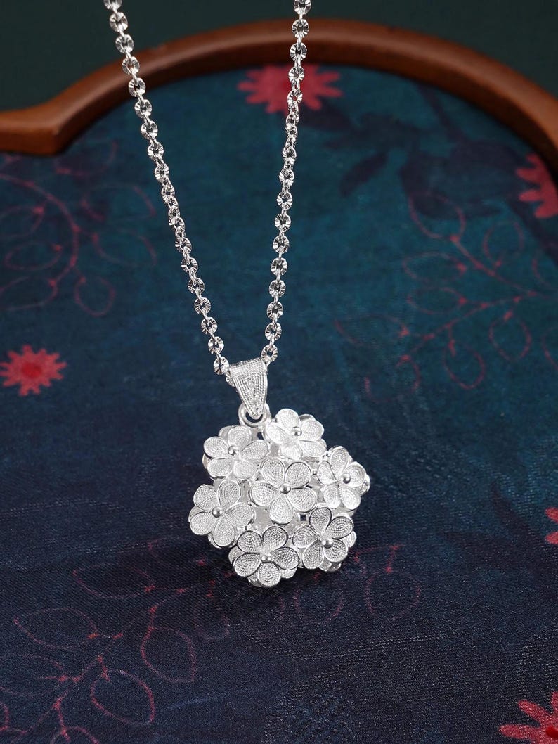 May include: A silver necklace featuring a cluster of small, detailed flower pendants. The pendant is suspended from a delicate chain. The necklace is displayed against a dark, patterned background.