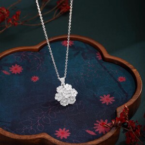 May include: A silver necklace with a floral pendant. The pendant is a cluster of small, detailed flowers. The necklace is displayed on a dark blue surface with red floral patterns, inside a wooden tray. Red floral accents are in the background.