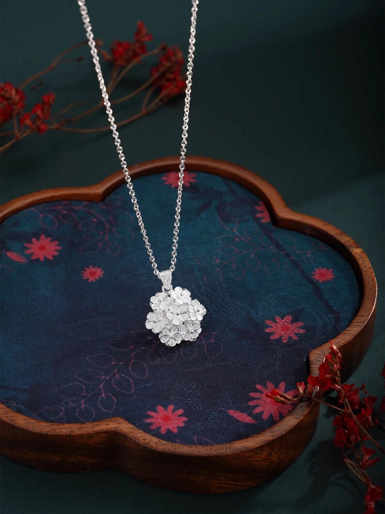 May include: A silver necklace with a floral pendant. The pendant is a cluster of small, detailed flowers. The necklace is displayed on a dark blue surface with red floral patterns, inside a wooden tray. Red floral accents are in the background.
