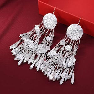 Könnte beinhalten: Ein Paar silberne Ohrringe mit filigranen Designs. Jeder Ohrring hat einen großen runden Anhänger mit einem kleineren runden Anhänger, der daran hängt. Die Anhänger sind durch eine Kette mit kleineren dekorativen Elementen verbunden.