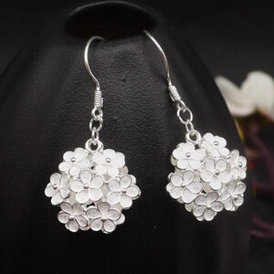 May include: A pair of silver-colored dangle earrings. Each earring features a cluster of small, detailed flowers. The earrings hang from a simple hook and are displayed against a dark background.