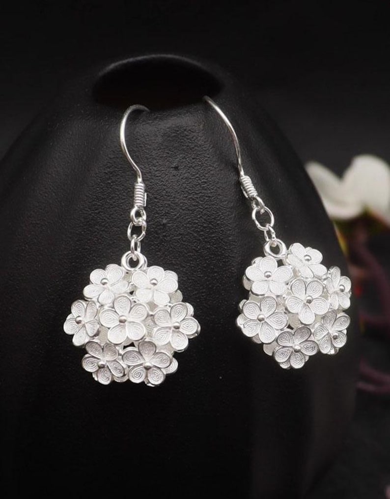 May include: A pair of silver-colored dangle earrings. Each earring features a cluster of small, detailed flowers. The earrings hang from a simple hook and are displayed against a dark background.