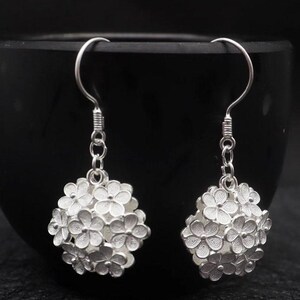 May include: A pair of silver-colored earrings. Each earring features a cluster of small, detailed flowers forming a spherical shape. The earrings hang from a simple hook design, with a textured section near the top.