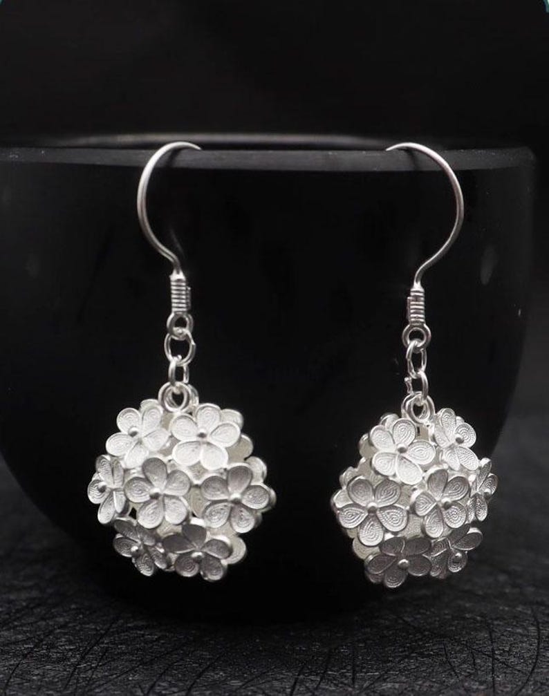 May include: A pair of silver-colored earrings. Each earring features a cluster of small, detailed flowers forming a spherical shape. The earrings hang from a simple hook design, with a textured section near the top.