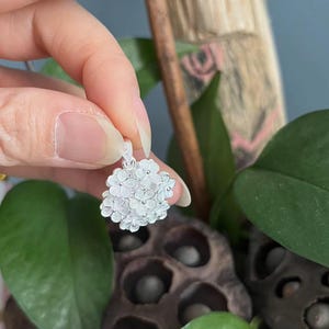 May include: A silver pendant featuring a cluster of small, detailed flowers. The pendant is held up against a backdrop of green leaves and a dark, textured surface. The floral design is intricate and delicate.