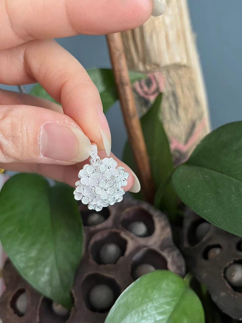 May include: A silver pendant featuring a cluster of small, detailed flowers. The pendant is held up against a backdrop of green leaves and a dark, textured surface. The floral design is intricate and delicate.