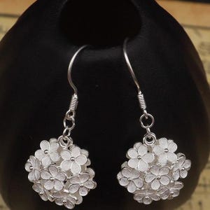 May include: A pair of silver-colored drop earrings. Each earring features a cluster of small, detailed flowers. The earrings are suspended from a simple hook and chain design, showcasing a delicate floral theme.