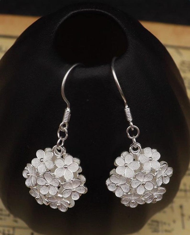 May include: A pair of silver-colored drop earrings. Each earring features a cluster of small, detailed flowers. The earrings are suspended from a simple hook and chain design, showcasing a delicate floral theme.