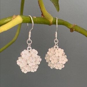 May include: A pair of silver-colored dangle earrings. Each earring features a cluster of small, detailed flowers, suspended from a simple hook. The earrings are displayed against a muted green and gray background, highlighting the delicate floral design.