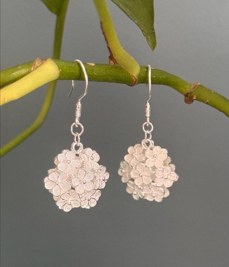 May include: A pair of silver-colored dangle earrings. Each earring features a cluster of small, detailed flowers, suspended from a simple hook. The earrings are displayed against a muted green and gray background, highlighting the delicate floral design.