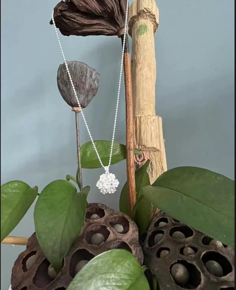 May include: A silver necklace with a round, clustered pendant. The pendant is made of small, reflective beads. The necklace hangs against a backdrop of green leaves, dried seed pods, and a wooden decorative element.