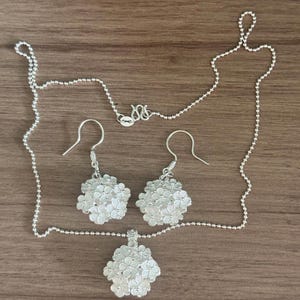 May include: A silver jewelry set featuring a necklace with a pendant and a pair of earrings. The pendant and earrings have a floral design. The necklace and earrings are made of silver and are displayed on a wooden surface.