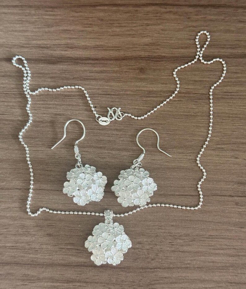 May include: A silver jewelry set featuring a necklace with a pendant and a pair of earrings. The pendant and earrings have a floral design. The necklace and earrings are made of silver and are displayed on a wooden surface.