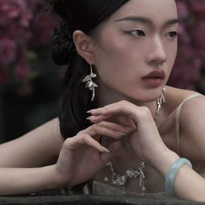 Può includere: Una donna indossa orecchini floreali argentati, una collana abbinata e un braccialetto verde chiaro. Gli orecchini e la collana presentano design delicati e pendenti. La donna ha i capelli scuri e indossa un top di colore chiaro.
