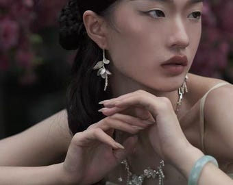 May include: A woman wearing silver floral earrings, a matching necklace, and a light green bracelet. The earrings and necklace feature delicate, dangling designs. The woman has dark hair and is wearing a light-colored top.