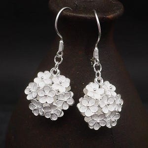 May include: A pair of silver-colored drop earrings. Each earring features a cluster of small, detailed flower shapes, creating a spherical design. The earrings hang from a simple hook and chain. The background is a dark, blurred setting.