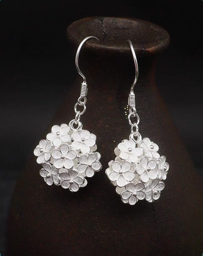 May include: A pair of silver-colored drop earrings. Each earring features a cluster of small, detailed flower shapes, creating a spherical design. The earrings hang from a simple hook and chain. The background is a dark, blurred setting.