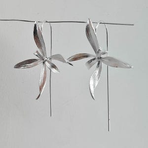 Könnte beinhalten: Ein Paar silberfarbene Ohrringe in Blumenform. Jeder Ohrring hat ein offenes Blumendesign mit mehreren Blütenblättern und einem langen, schlanken Stift. Die Ohrringe sind an einem dünnen Draht aufgehängt.