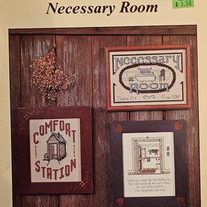 May include: A collection of framed cross-stitch samplers with bathroom-themed designs. The samplers are displayed on a wooden background. One sampler reads "Necessary Room," another "Comfort Station," and a third features a bathroom scene with text.
