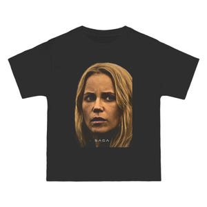 May include: Black t-shirt with a portrait of a woman with blonde hair and a serious expression. The word "SAGA" is printed in white at the bottom. The t-shirt is made of a soft material and is suitable for casual wear.