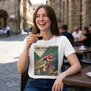 May include: White t-shirt featuring a vintage-style graphic of a cyclist in a Giro d'Italia race. The design includes the text "Giro D'Italia 2026" and race details. The woman is smiling and holding a cup of coffee.