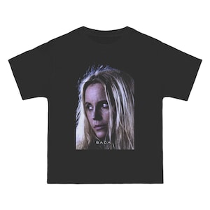 May include: Black t-shirt featuring a portrait of a woman with long blonde hair. The image is centered on the front of the shirt, with the word "SAGA" printed below the portrait. The t-shirt has short sleeves and a crew neck.