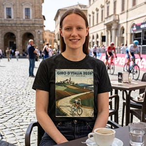 May include: A woman wearing a black t-shirt featuring a vintage-style poster for the Giro d'Italia 2026 cycling race. The poster depicts a cyclist on a winding road through a rural landscape with rolling hills and fields.