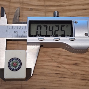 May include: A rectangular off-white Mahjong tile featuring a circular floral design in blue, green, and red. The tile is positioned within a silver digital caliper showing a measurement of 0.7425 inches on its clear liquid crystal display screen.