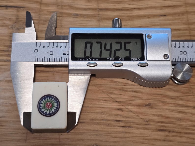 May include: A rectangular off-white Mahjong tile featuring a circular floral design in blue, green, and red. The tile is positioned within a silver digital caliper showing a measurement of 0.7425 inches on its clear liquid crystal display screen.