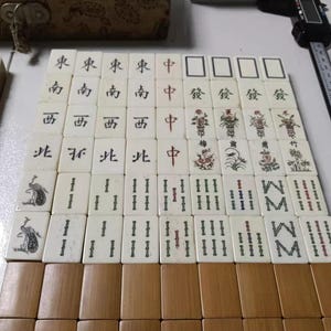 Puede incluir: Un conjunto de fichas de Mahjong de color blanco hueso dispuestas en cuadr&iacute;cula, con caracteres en tinta negra y roja, ilustraciones florales y motivos de aves. Las fichas se muestran junto a un calibre digital y una caja de madera.