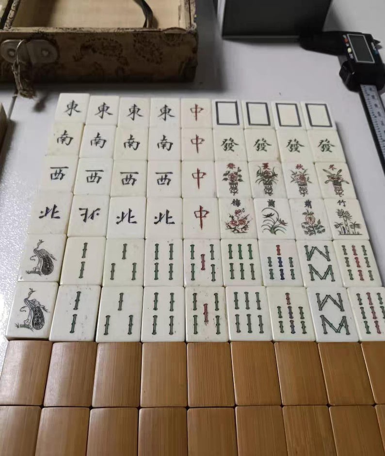 Puede incluir: Un conjunto de fichas de Mahjong de color blanco hueso dispuestas en cuadr&iacute;cula, con caracteres en tinta negra y roja, ilustraciones florales y motivos de aves. Las fichas se muestran junto a un calibre digital y una caja de madera.