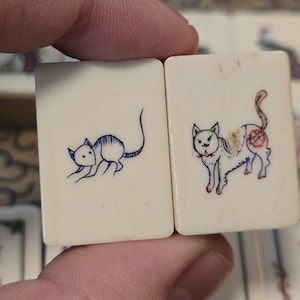 May include: Two rectangular off-white tiles featuring hand-engraved illustrations. The left tile displays a blue ink drawing of a mouse, while the right tile features a multi-colored ink drawing of a cat with red, blue, and yellow details.
