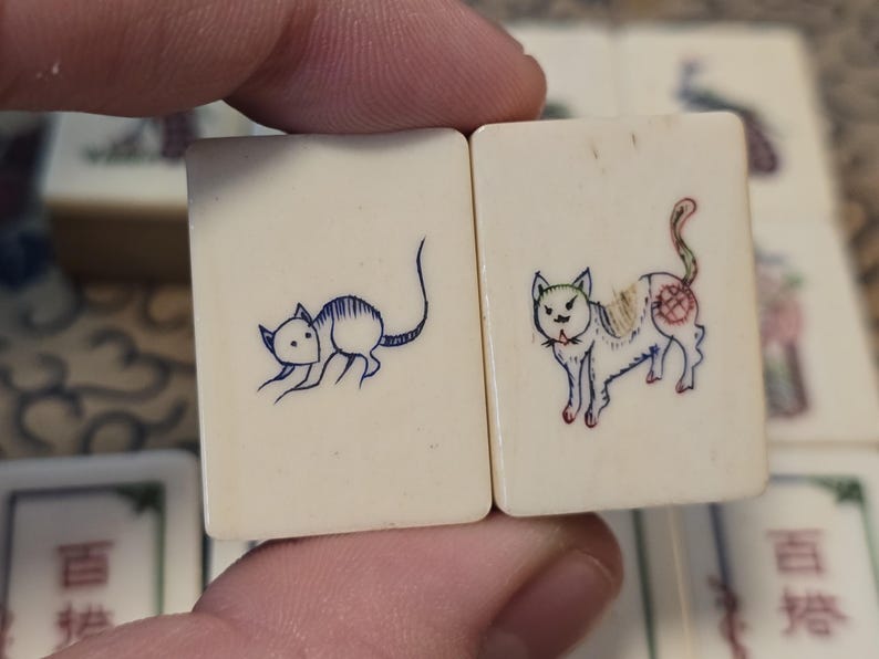 May include: Two rectangular off-white tiles featuring hand-engraved illustrations. The left tile displays a blue ink drawing of a mouse, while the right tile features a multi-colored ink drawing of a cat with red, blue, and yellow details.