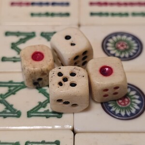 May include: Four off-white dice with weathered textures resting on a grid of mahjong tiles. The dice feature black and red pips, with some faces showing single red circular indentations. The background tiles display green characters and circular floral patterns.