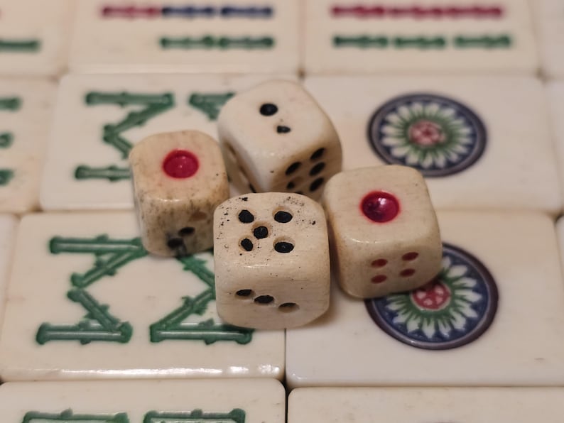May include: Four off-white dice with weathered textures resting on a grid of mahjong tiles. The dice feature black and red pips, with some faces showing single red circular indentations. The background tiles display green characters and circular floral patterns.