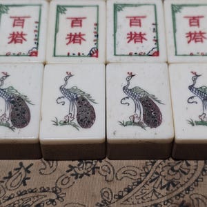 Puede incluir: Ocho fichas de mahjong rectangulares dispuestas en dos filas. La fila superior presenta cuatro fichas con caracteres chinos en rojo y verde. La fila inferior muestra cuatro fichas, cada una decorada con un pavo real ilustrado de perfil.