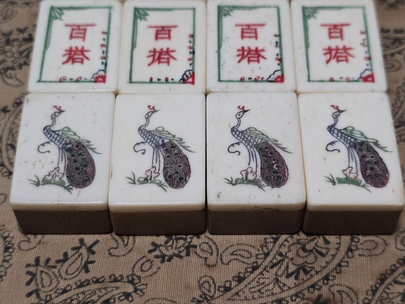 Puede incluir: Ocho fichas de mahjong rectangulares dispuestas en dos filas. La fila superior presenta cuatro fichas con caracteres chinos en rojo y verde. La fila inferior muestra cuatro fichas, cada una decorada con un pavo real ilustrado de perfil.