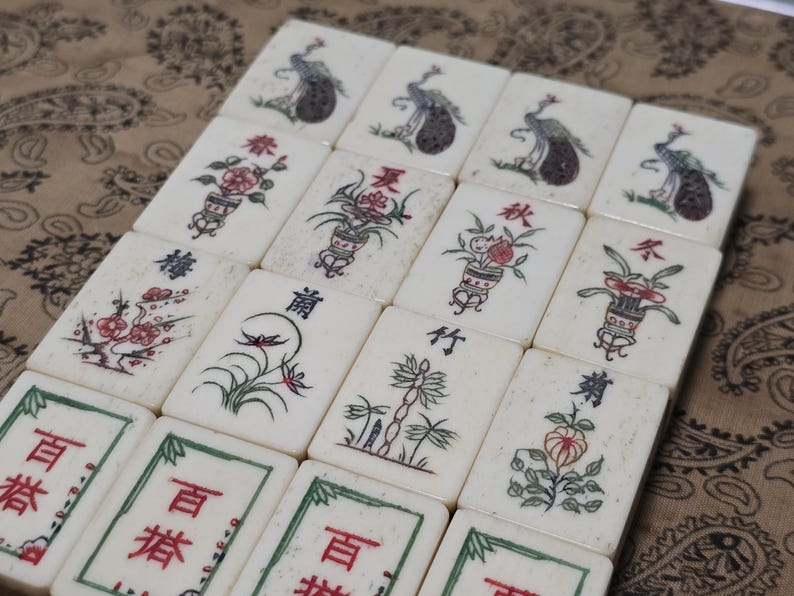 Puede incluir: Una colecci&oacute;n de fichas de Mahjong rectangulares de color blanco roto dispuestas en una cuadr&iacute;cula. Las fichas presentan ilustraciones pintadas a mano de pavos reales, arreglos florales y motivos bot&aacute;nicos en rojo, verde y negro, junto con caracteres chinos.