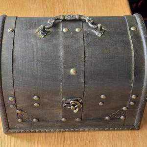 Puede incluir: Un cofre de almacenamiento de color marr&oacute;n oscuro con tapa arqueada y acabado desgastado. Presenta un asa met&aacute;lica decorativa en la tapa, un cierre central de metal y tachuelas de color lat&oacute;n a lo largo de los bordes y las esquinas.