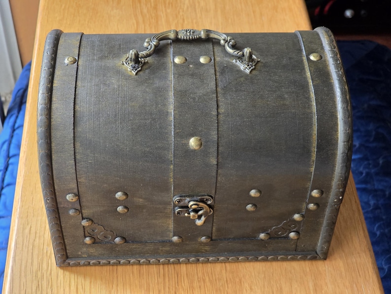 Puede incluir: Un cofre de almacenamiento de color marr&oacute;n oscuro con tapa arqueada y acabado desgastado. Presenta un asa met&aacute;lica decorativa en la tapa, un cierre central de metal y tachuelas de color lat&oacute;n a lo largo de los bordes y las esquinas.