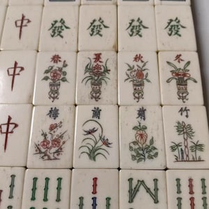 Puede incluir: Una colecci&oacute;n de fichas de mahjong rectangulares de color blanco hueso dispuestas en una cuadr&iacute;cula. Las fichas presentan varios dise&ntilde;os grabados y pintados, incluyendo caracteres chinos rojos, motivos florales verdes y plantas.