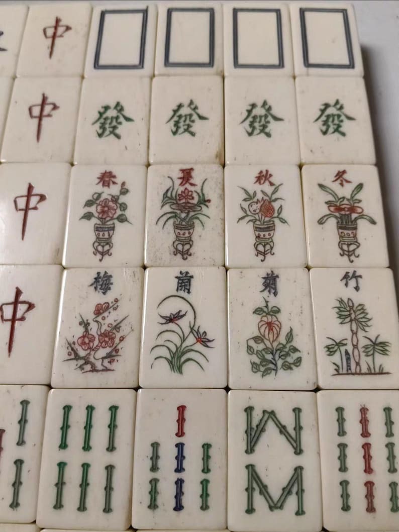Puede incluir: Una colecci&oacute;n de fichas de mahjong rectangulares de color blanco hueso dispuestas en una cuadr&iacute;cula. Las fichas presentan varios dise&ntilde;os grabados y pintados, incluyendo caracteres chinos rojos, motivos florales verdes y plantas.