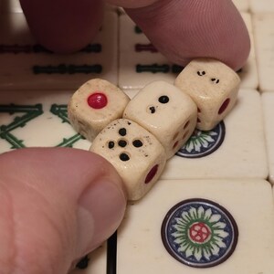 May include: Three small, off-white, aged-looking dice with black pips and red circular inlays, positioned on top of a set of cream-colored mahjong tiles featuring green and blue floral designs and black character markings.