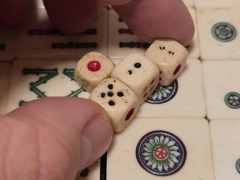 May include: Three small, off-white, aged-looking dice with black pips and red circular inlays, positioned on top of a set of cream-colored mahjong tiles featuring green and blue floral designs and black character markings.