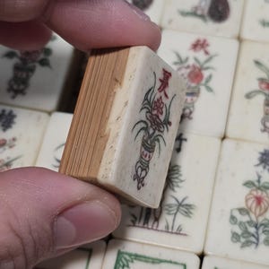 Puede incluir: Una ficha de Mahjong rectangular con un frente de color crema que presenta un car&aacute;cter chino rojo y una ilustraci&oacute;n floral verde. La parte posterior de la ficha est&aacute; hecha de madera de color marr&oacute;n claro con veta vertical visible.