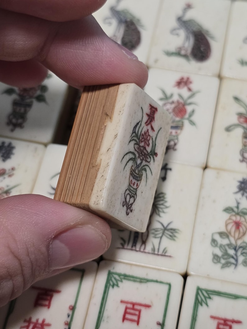 Puede incluir: Una ficha de Mahjong rectangular con un frente de color crema que presenta un car&aacute;cter chino rojo y una ilustraci&oacute;n floral verde. La parte posterior de la ficha est&aacute; hecha de madera de color marr&oacute;n claro con veta vertical visible.
