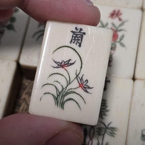 Puede incluir: Una ficha de Mahjong rectangular de color blanco hueso con un dise&ntilde;o floral pintado a mano en verde y rojo de una orqu&iacute;dea con hojas largas y curvas. La parte superior de la ficha muestra un car&aacute;cter chino negro y una textura veteada.