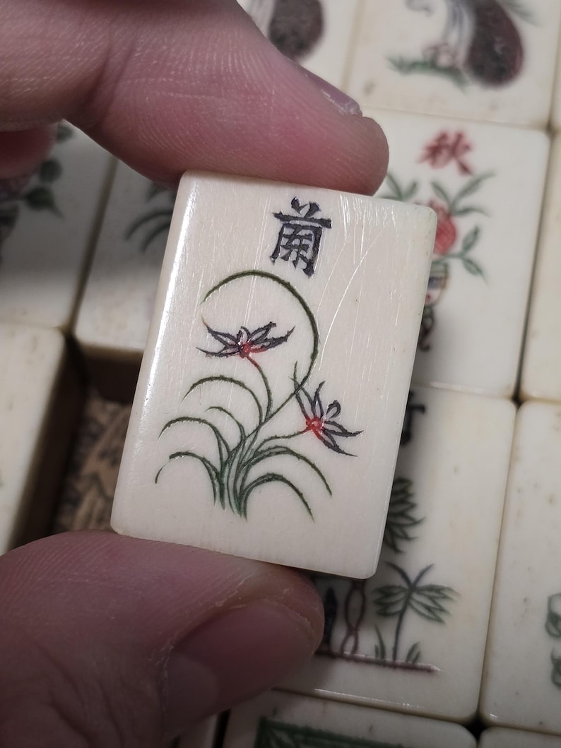 Puede incluir: Una ficha de Mahjong rectangular de color blanco hueso con un dise&ntilde;o floral pintado a mano en verde y rojo de una orqu&iacute;dea con hojas largas y curvas. La parte superior de la ficha muestra un car&aacute;cter chino negro y una textura veteada.