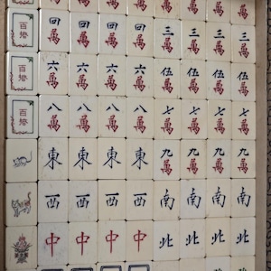 May include: A set of off-white Mahjong tiles arranged in a grid within a box. The tiles feature various Chinese characters in red, blue, and green ink, along with illustrations of animals and figures on the left column of tiles.