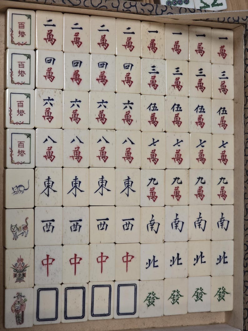 May include: A set of off-white Mahjong tiles arranged in a grid within a box. The tiles feature various Chinese characters in red, blue, and green ink, along with illustrations of animals and figures on the left column of tiles.